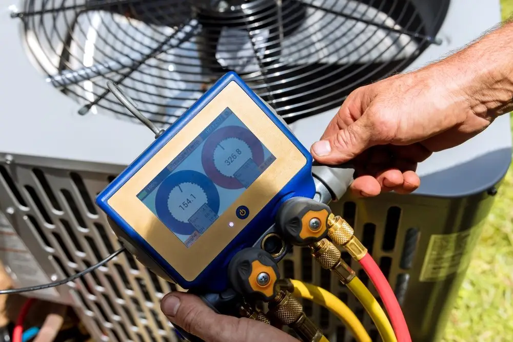 A person holding a digital manifold gauge with yellow and red hoses attached, used for HVAC or refrigeration systems, near an outdoor air conditioning unit.