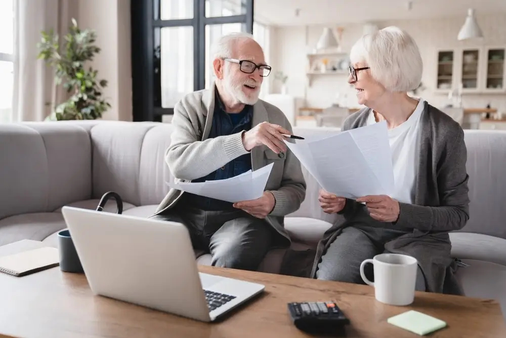 alt="An elderly man and woman are sitting on a light-colored sofa in a bright living room, smiling and discussing papers with a laptop, coffee mug, and calculator on the table in front of them."