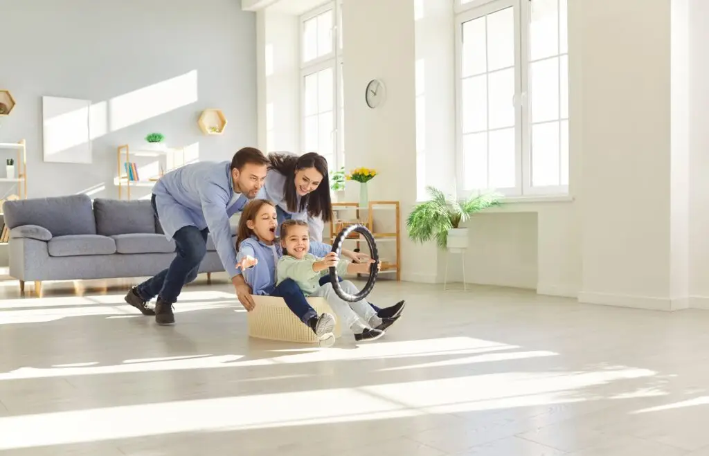 A family of four enjoying playtime indoors. The father, mother, and two young girls are riding a cardboard box turned into a toy car, with the girls holding a steering wheel attached to the box. They are smiling and laughing in a bright, spacious living room with large windows, plants, and minimal decor.
